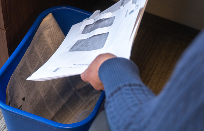 A person putting mail and a newspaper in a recycling bin.