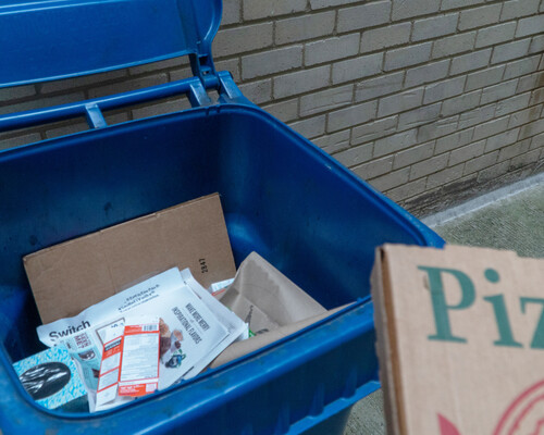 A pizza box being placed in a blue recycling cart already filled with paper items.