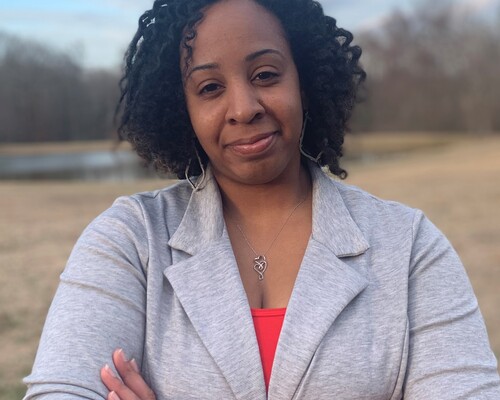 Alondria Hamilton stands with her arms crossed while smiling at the camera. There are trees in the background.