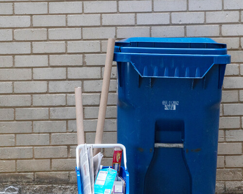 A paper recycling cart and a small recycling container that has paper recyclables in it outside of a brick building.