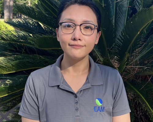 Hong Wilcoxon is wearing glasses and a polo. She's standing outside in front of plants.