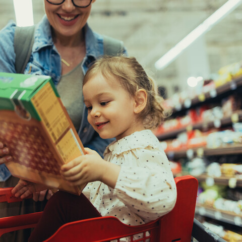 A child smiles while holding a cereal box as she sits in a grocery cart. Her mother smiles in the background.